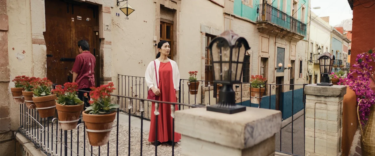 Angel Flight THE MOVIE (2026) movie still: fullbody — A narrow cobblestone street lined with beige stucco buildings and iron balcony railings. A…