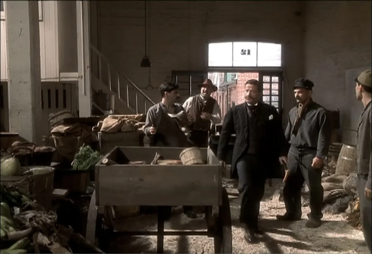 Vendetta (2022) movie still: fullbody — A rustic warehouse interior contains wooden carts, wooden crates, and sacks of produce. Four men dressed in…