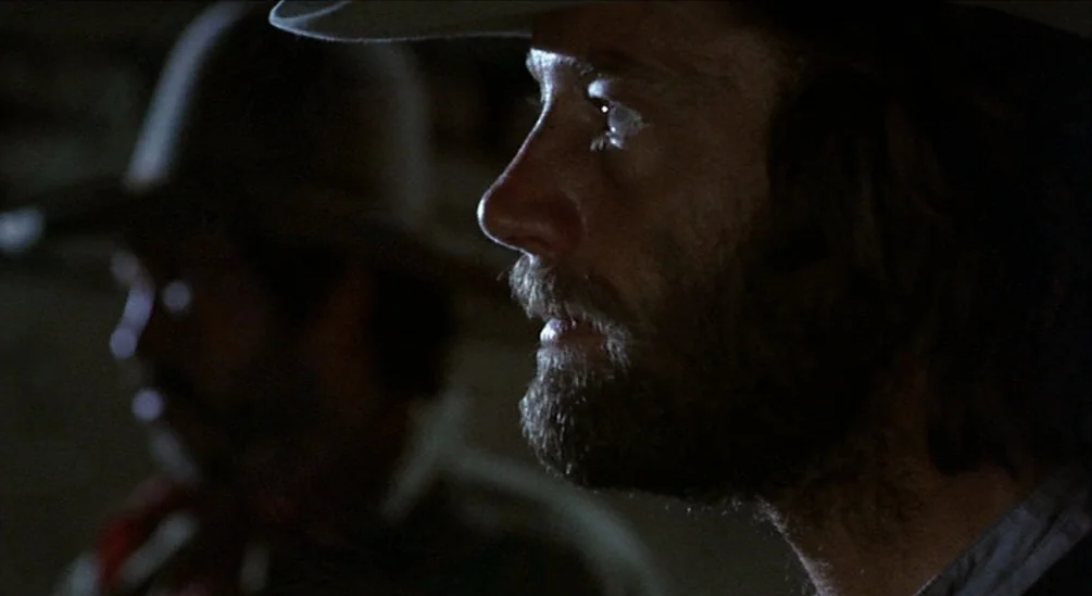 The Hired Hand (1971) movie still: closeup — A dark, dimly lit interior space with minimal architectural detail visible in the shadows. A man with a…