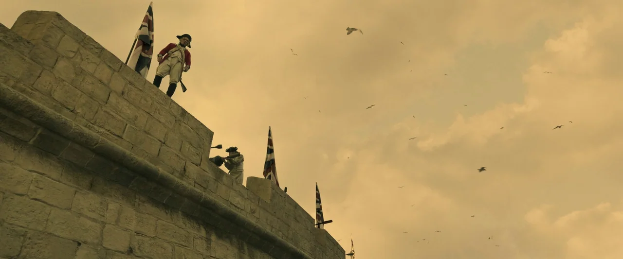 Napoleon (2023) movie still: wide — A stone fortification wall rises against an expansive sky filled with flying birds. Two soldiers in period…