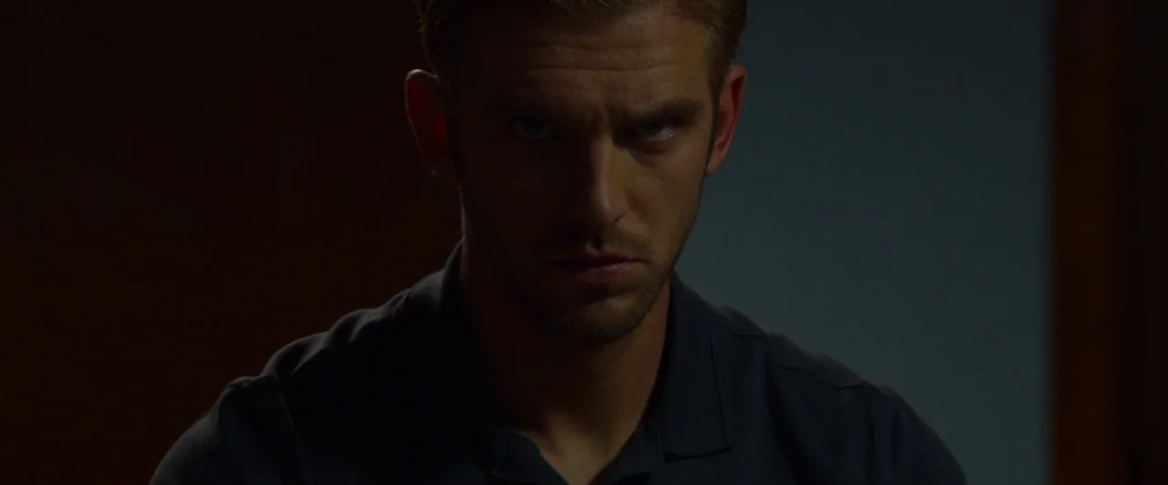 The Guest (2014) movie still: closeup — An interior space is dimly lit with significant shadows obscuring much of the environment. A man in a dark…