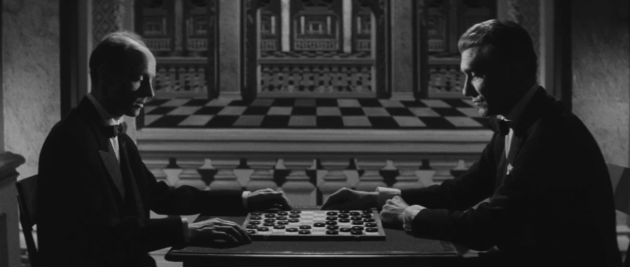 Last Year at Marienbad (1961) movie still: medium — Two men in tuxedos sit at a square table playing a game of checkers. The wall behind them…