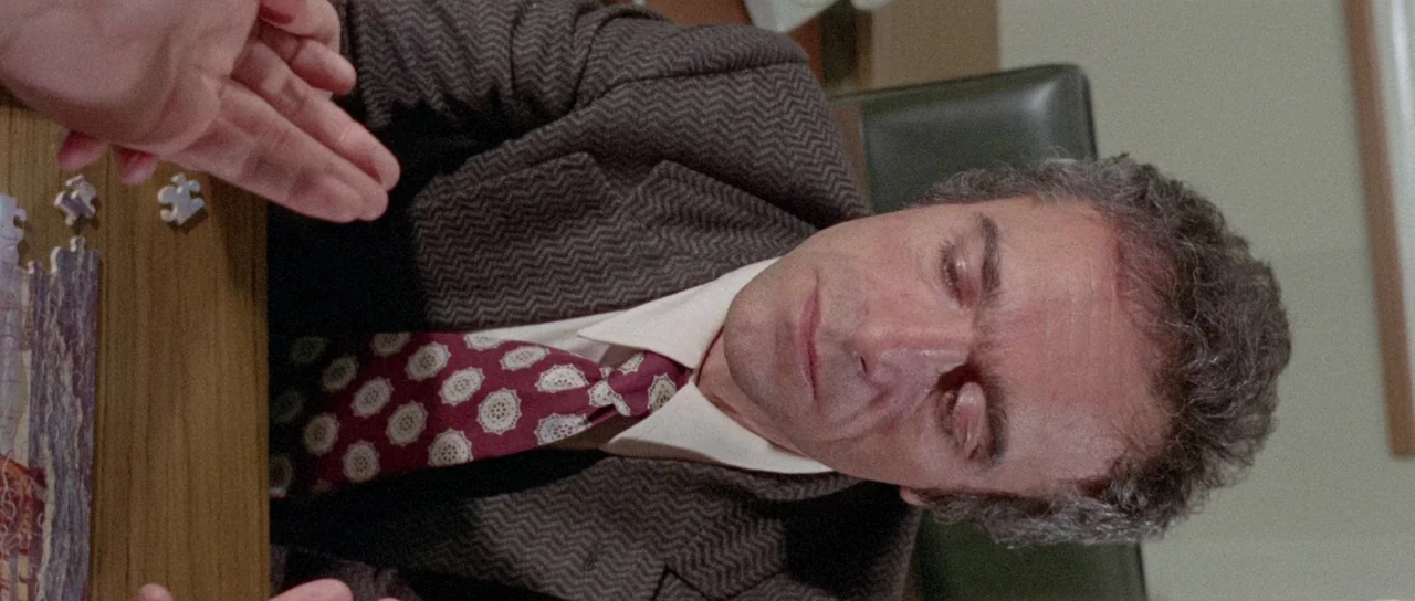 The Case of the Scorpion’s Tail (1971) movie still: medium — A man in a herringbone jacket sits at a wooden desk assembling a puzzle. His focus is…