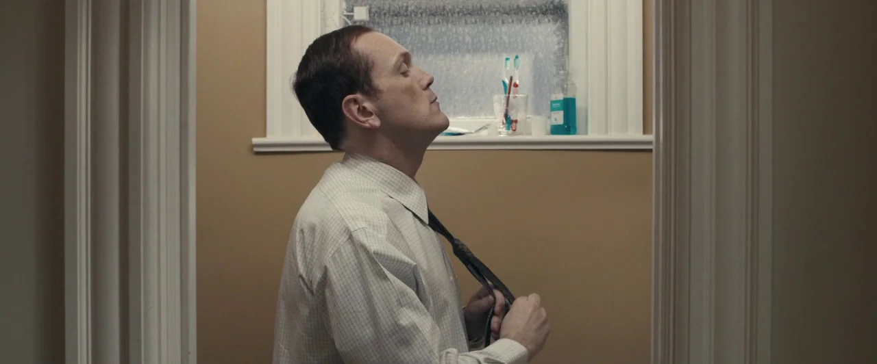 Compliance (2012) movie still: medium — A man stands in a brightly lit, beige-walled space near a frosted window. He holds his dark necktie with…