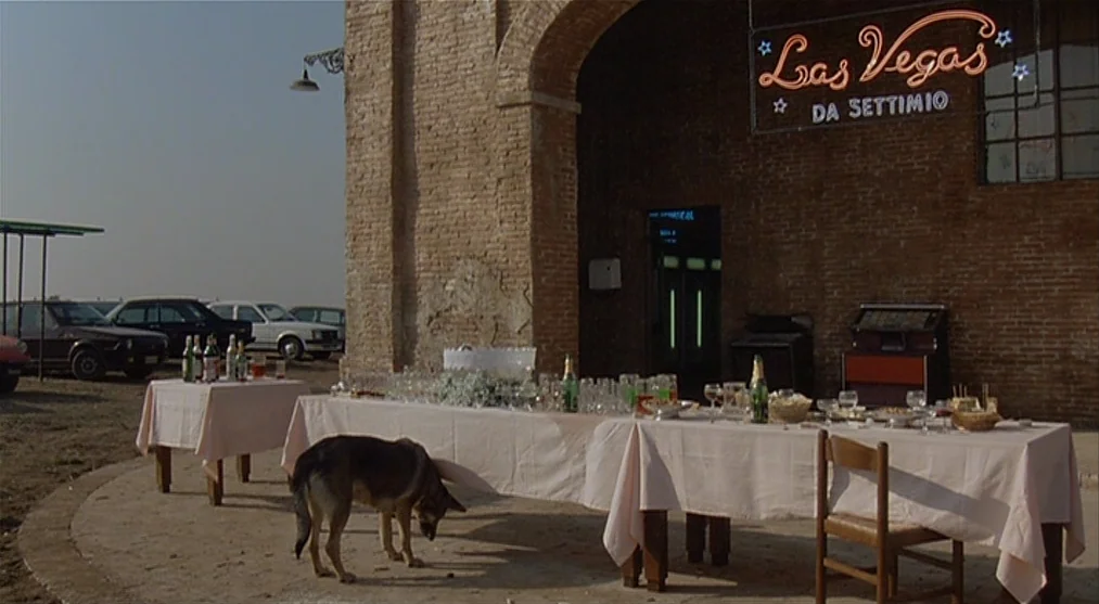 The Voice of the Moon (1990) movie still: wide — An outdoor banquet setup sits before a brick building with a neon sign displayed. A german shepherd…