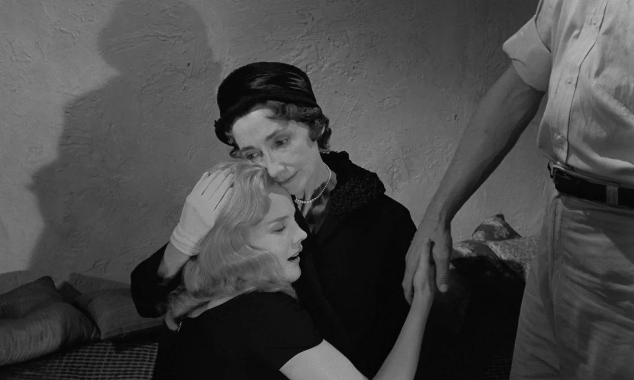 Something Wild (1961) movie still: medium — A dimly lit room with plain walls and soft pillows on a low bench. An older woman embraces a younger…