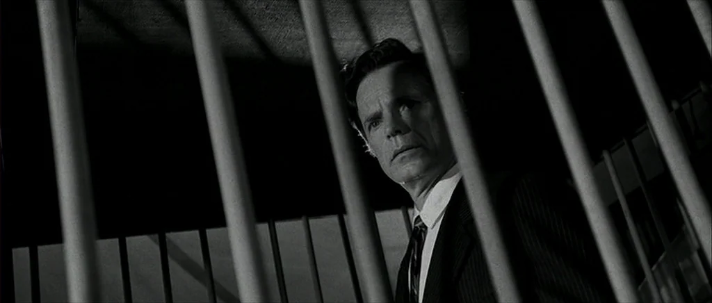 I’m Not There (2007) movie still: medium — A dark stairwell featuring thick, vertical metal bars in the foreground. A man in a suit looks toward the…