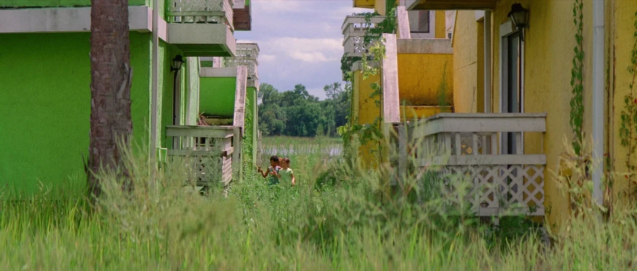 The Florida Project (2017) movie still: wide — A pair of colorful, stucco apartment buildings with white lattice balconies stand near a swamp. Two…