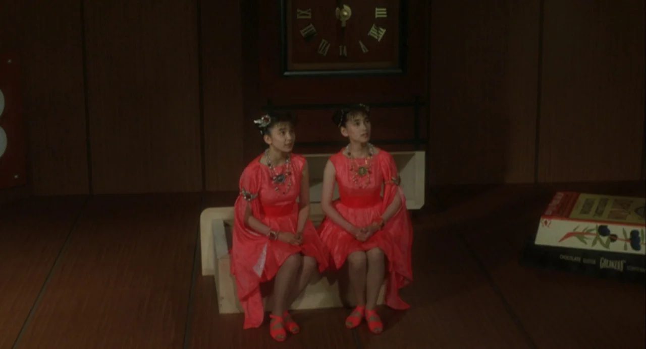 Godzilla and Mothra: The Battle for Earth (1992) movie still: medium — Two identical twin women in bright red dresses sit together on a small bench.…