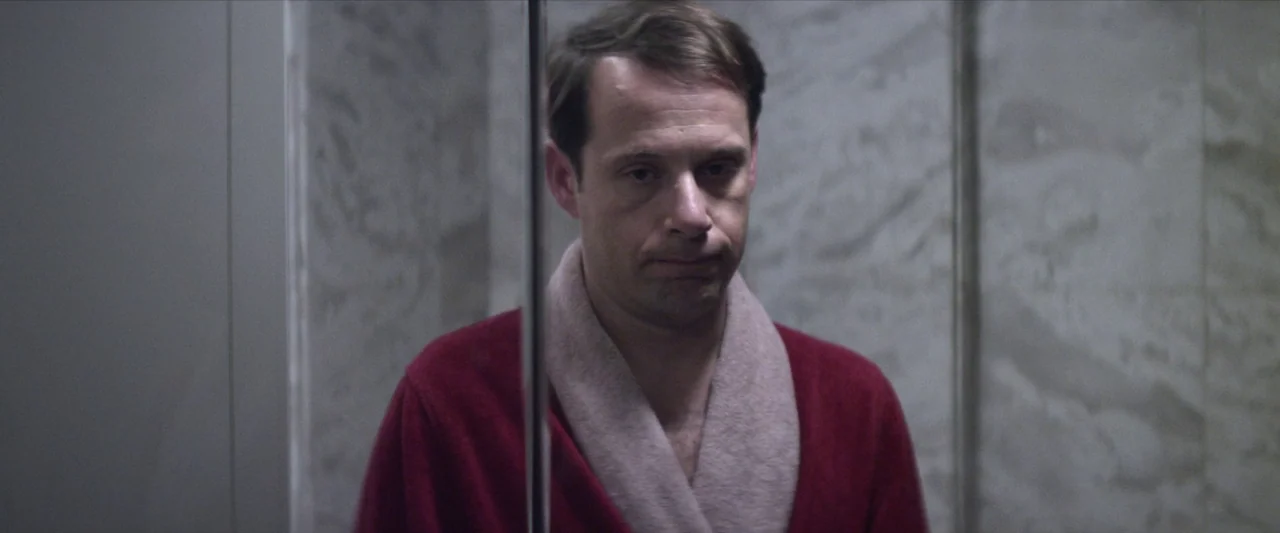 Chevalier (2015) movie still: closeup — A man looks into a bathroom mirror in a tiled space. The vertical edge of the mirror bisects the frame.
