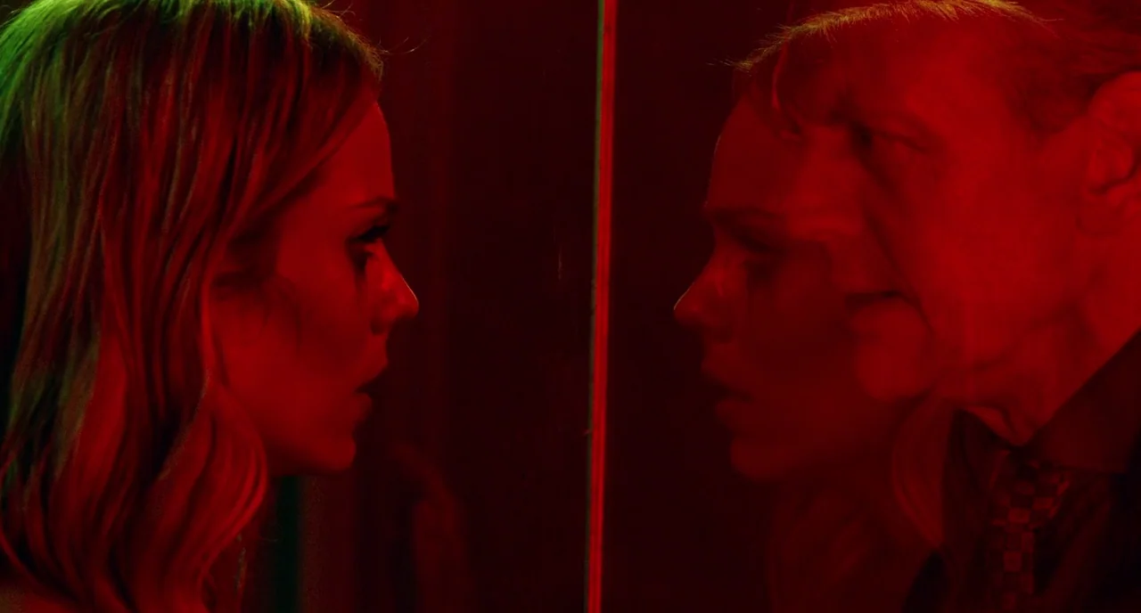 Rabid (2019) movie still: medium — A dark space features a large reflective surface casting a layered, mirrored image. A blonde woman faces an older…