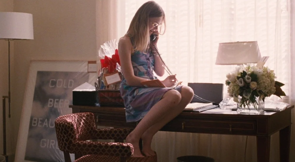 Somewhere (2010) movie still: medium — A sunlit room features a dark wood desk holding a telephone and floral arrangement. A young woman sits on the…