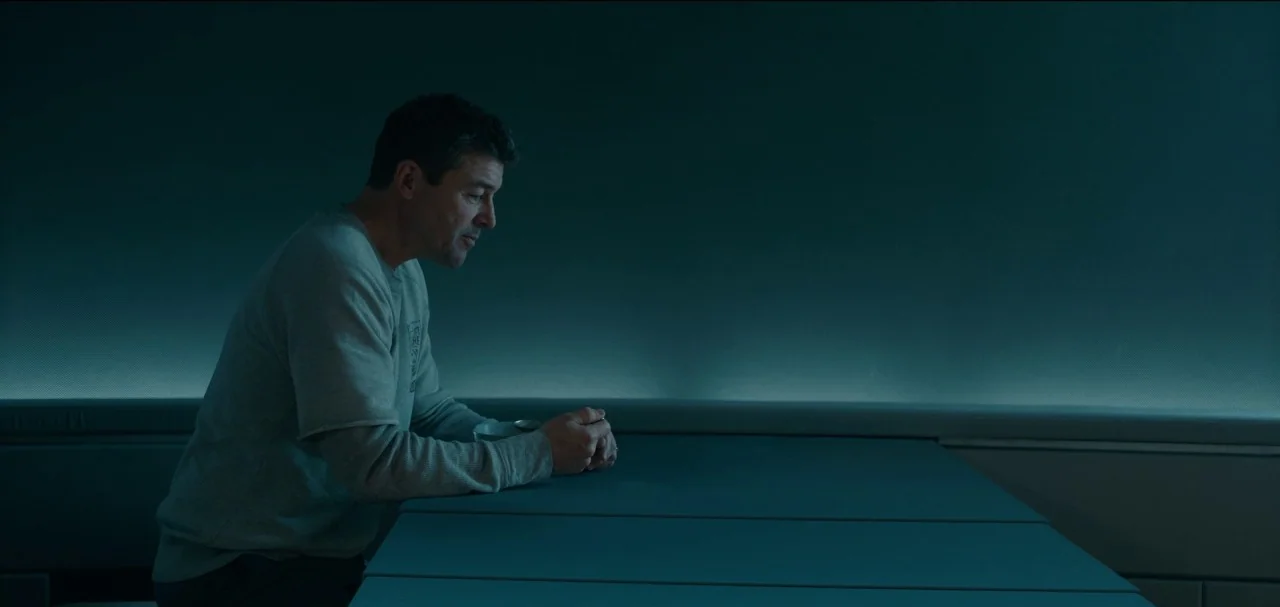 The Midnight Sky (2020) movie still: medium — A dark breakroom features a smooth, flat table surface and plain, dark walls. The man rests his hands…