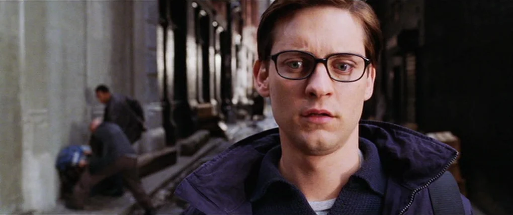 Spider-Man 2 (2004) movie still: closeup — An urban alleyway with dark buildings and blurred figures in the background. A young man stares directly…