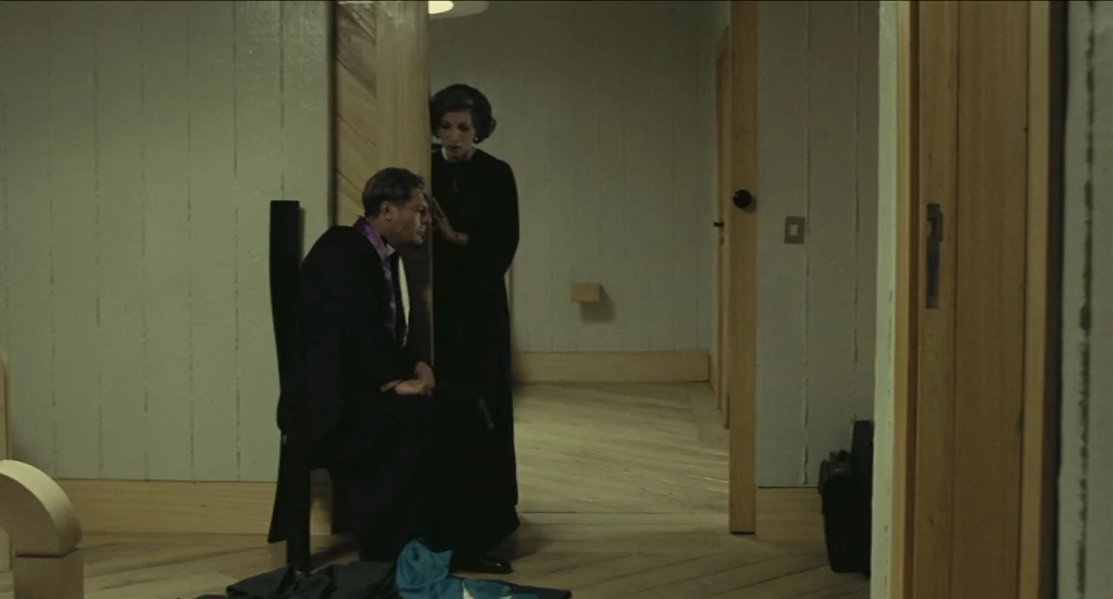 Todo Modo (1976) movie still: medium — A dimly lit modern hallway features light-colored wood paneling on walls and floor. A priest sits on a black…