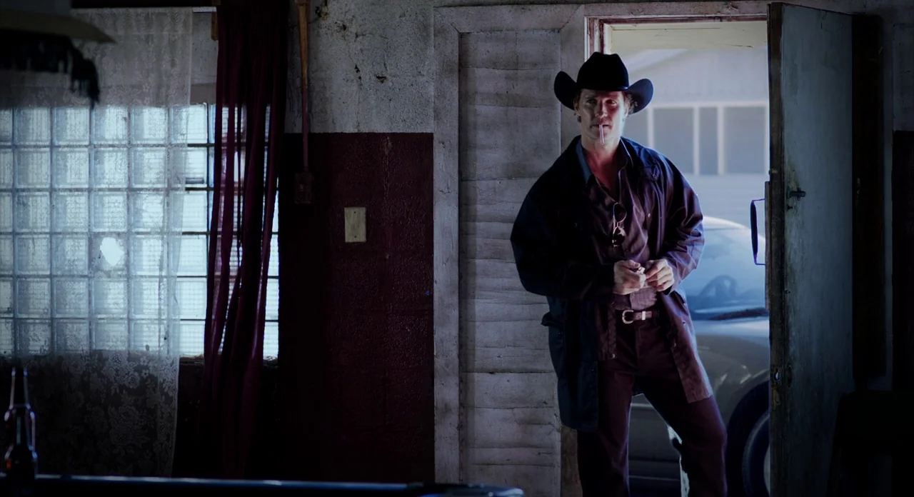 Killer Joe (2011) movie still: fullbody — A dimly lit room with peeling walls features an open door to daylight. A man in a cowboy hat stands in the…