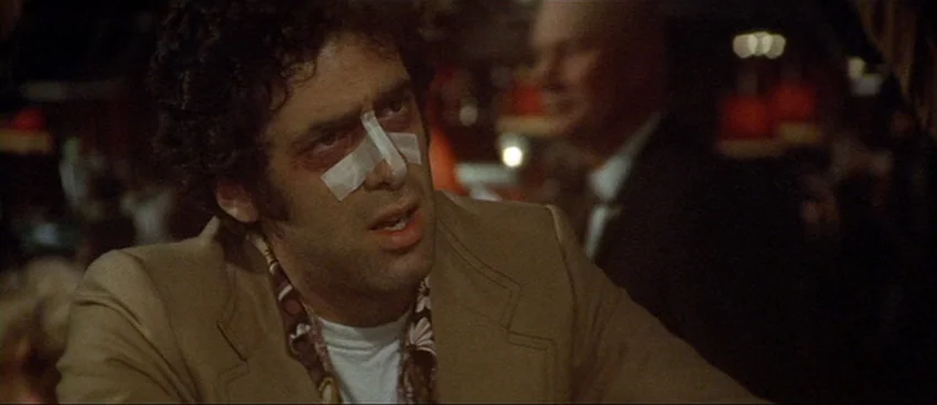 California Split (1974) movie still: medium — A dimly lit room with blurred silhouettes of people seated in the background. A man sits in the…