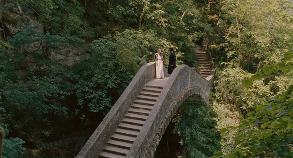 A Dangerous Method movie still: wide — A stone arched bridge crosses a ravine surrounded by dense green forest trees. A man and a woman in…