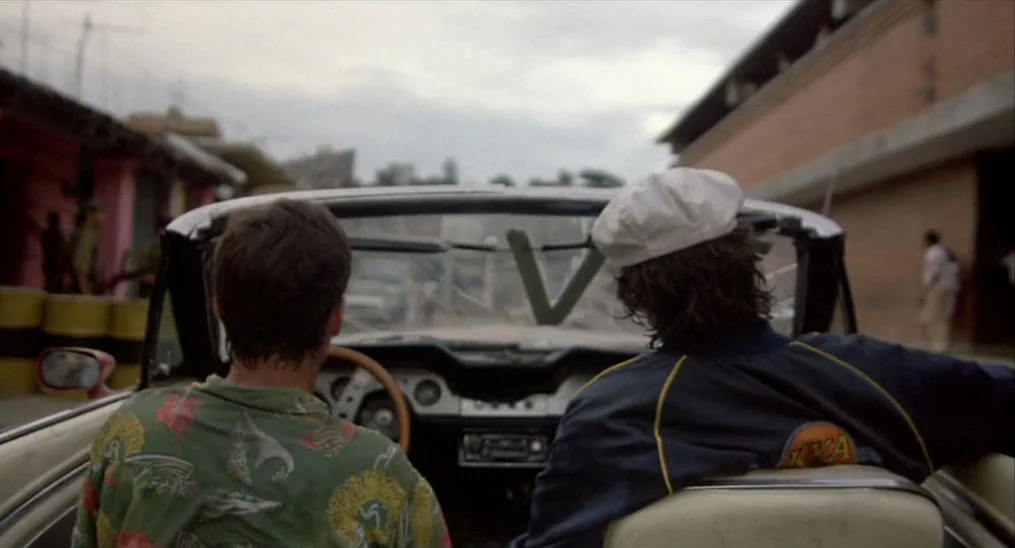 Salvador (1986) movie still: medium — A view from the back seat of a convertible car on an overcast day. Two men sit in the front seats while the…
