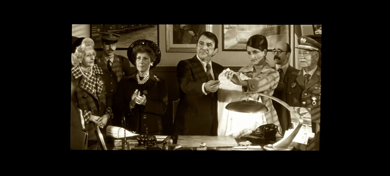 Live Flesh (1997) movie still: medium — An office interior features several people standing around a desk with a lamp. A man and a woman in the…