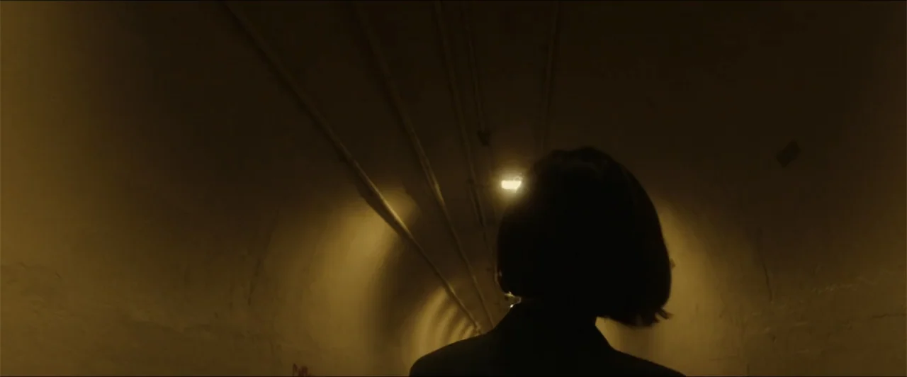 Exposed (2016) movie still: medium — A narrow arched concrete tunnel is illuminated by a single overhead yellow light fixture. A person with a short…