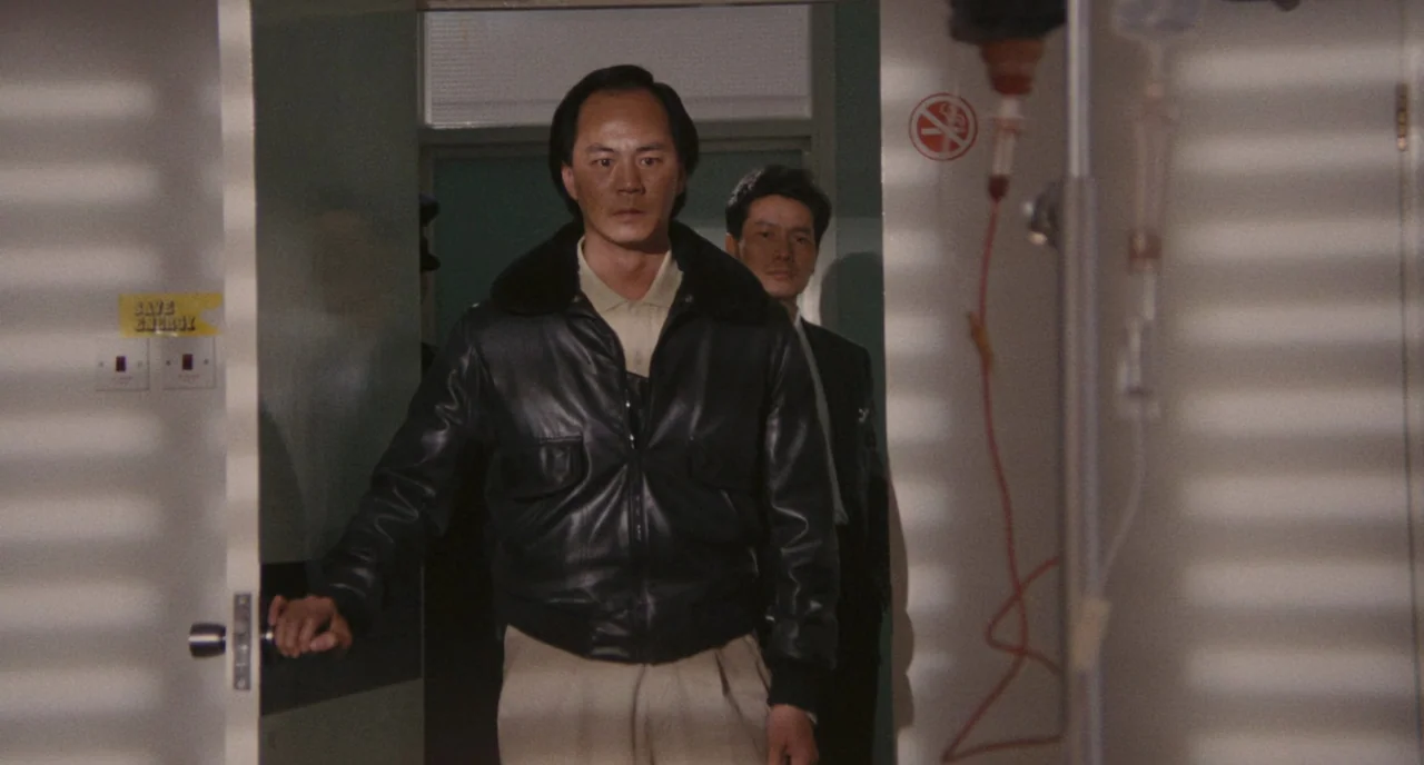 A Better Tomorrow (1986) movie still: medium — A hospital doorway features a cream-colored door and a no-smoking sign on the wall. Two men in formal…