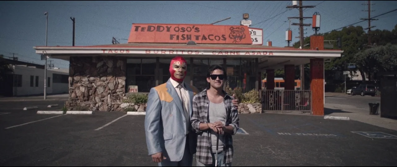 Lowlife (2017) movie still: fullbody — A roadside taco stand with a stone facade and orange signage stands at dusk. Two men stand in the paved…