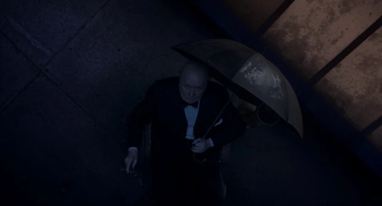 Darkest Hour (2017) movie still: medium — An elderly man in a dark suit sits on a paved outdoor area at night. He holds an umbrella overhead to…