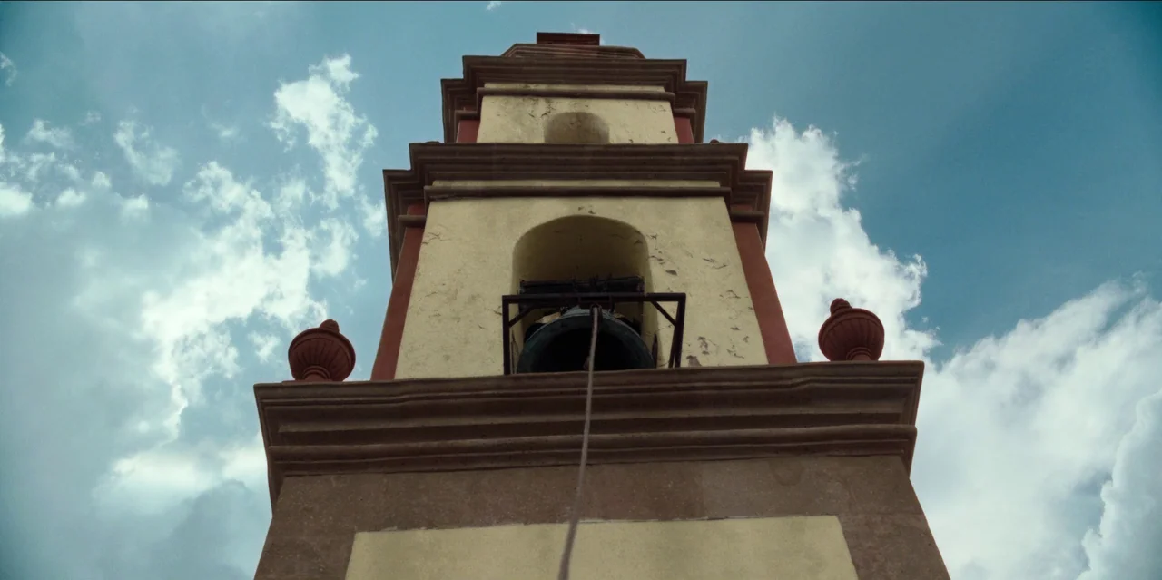 Pedro Páramo (2024) movie still: establishing — A stone bell tower features yellow and red masonry with a central hanging bell. The structure rises…