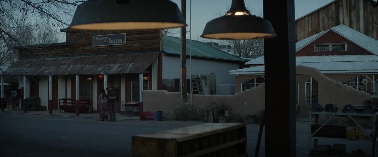Cry Macho (2021) movie still: wide — A quiet street in a small town features adobe architecture under a fading blue sky. A man and woman stand…