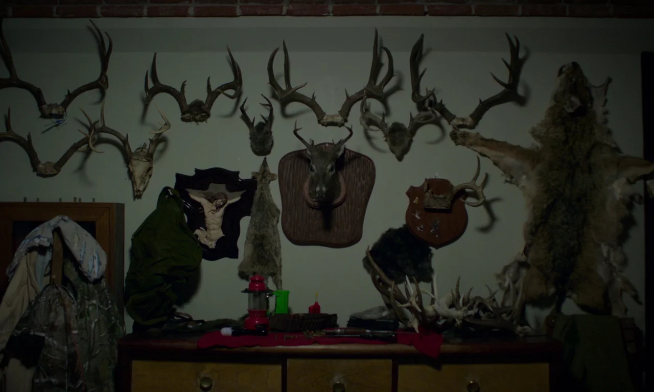 The Untamed (2016) movie still: medium — A wall decorated with numerous taxidermy deer antlers and a hanging fur pelt. A wooden table sits below,…