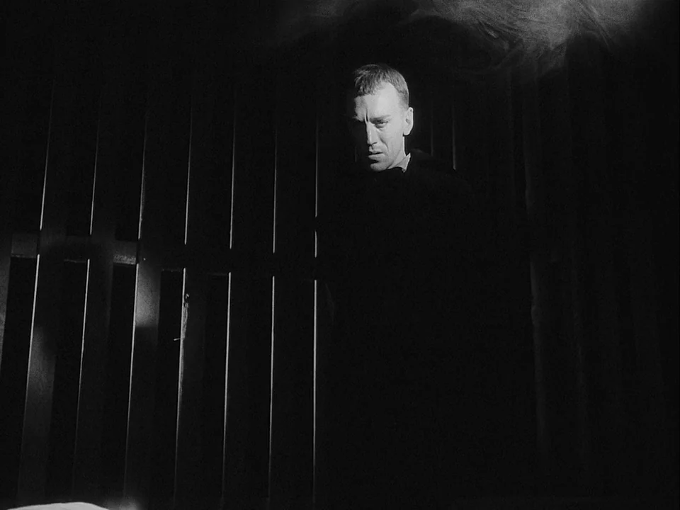 The Magician (1958) movie still: medium — A man stands before a dark vertical slatted gate in an interior space. He wears a dark coat and looks…