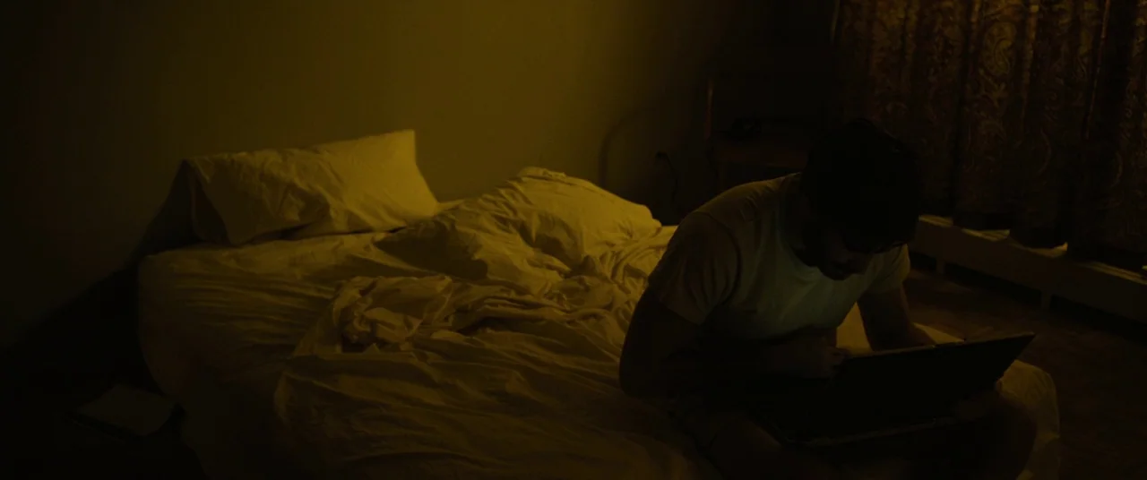 Enemy (2013) movie still: medium — A dark bedroom with an unmade bed featuring rumpled white sheets and pillows. A man sits on the edge of the…