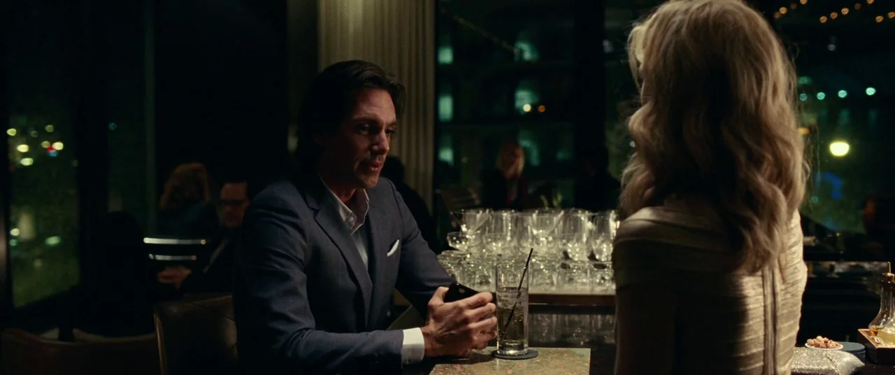 Widows (2018) movie still: medium — A dark upscale bar with patrons visible in the blurred background under warm lights. A man in a blue suit sits…