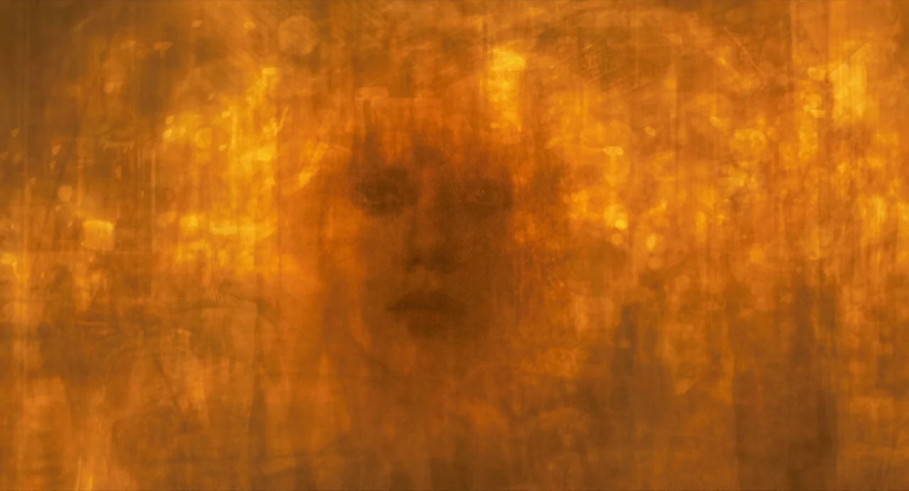 Under The Skin (2013) movie still: closeup — An abstract, textured orange filter obscures a human face. A woman looks directly toward the viewer.