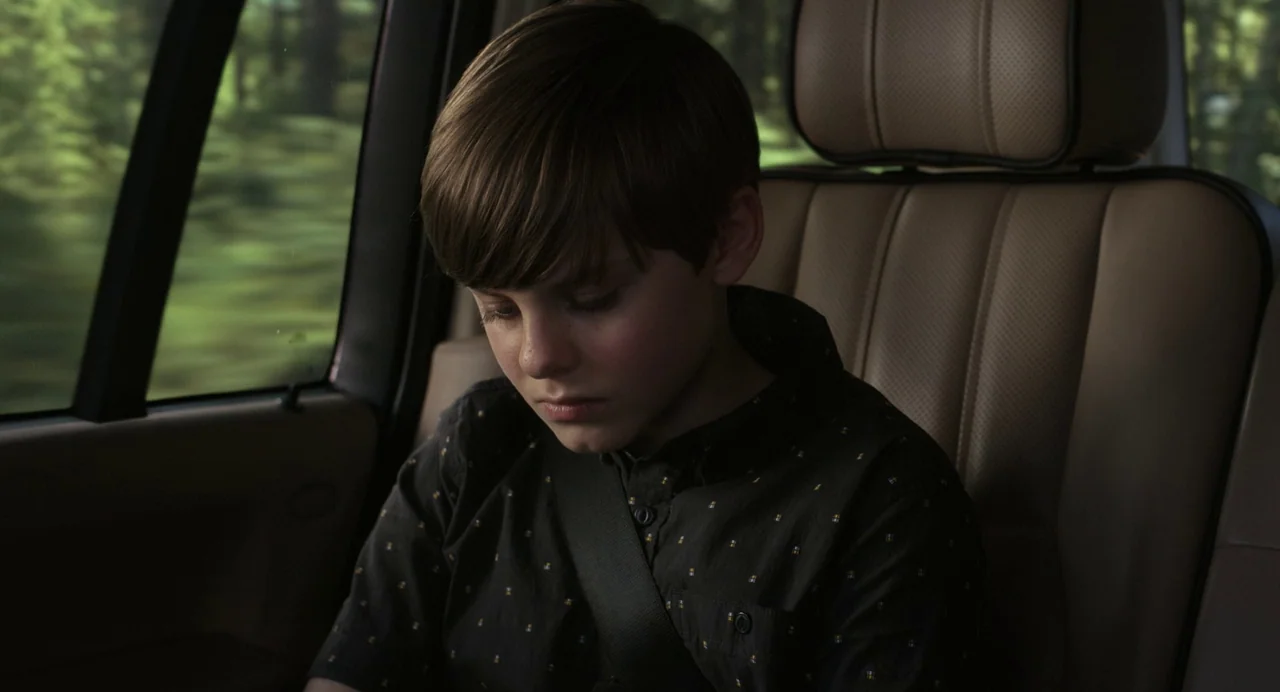 Goodnight Mommy (2022) movie still: medium — A young boy sits in the back of a moving car during the day. He stares downwards while the wooded…
