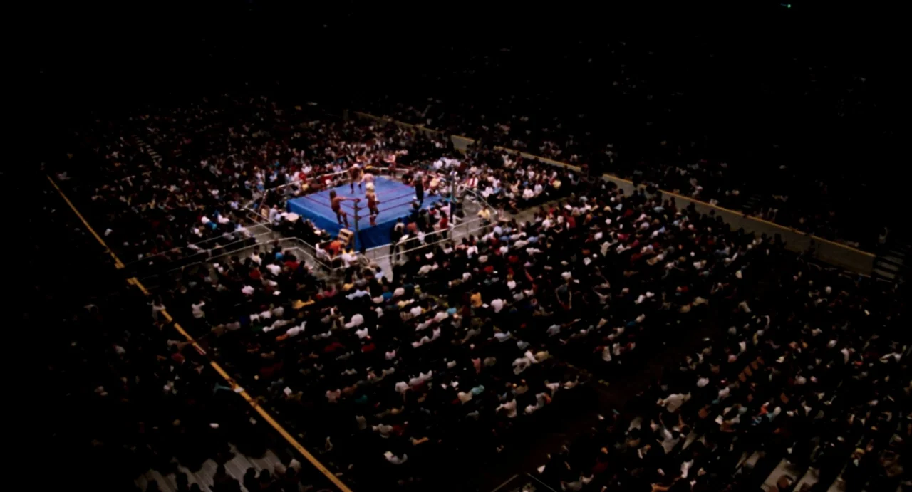 Highlander (1986) movie still: establishing — A large indoor arena is filled with a dense crowd of spectators in darkness. In the center, bright…