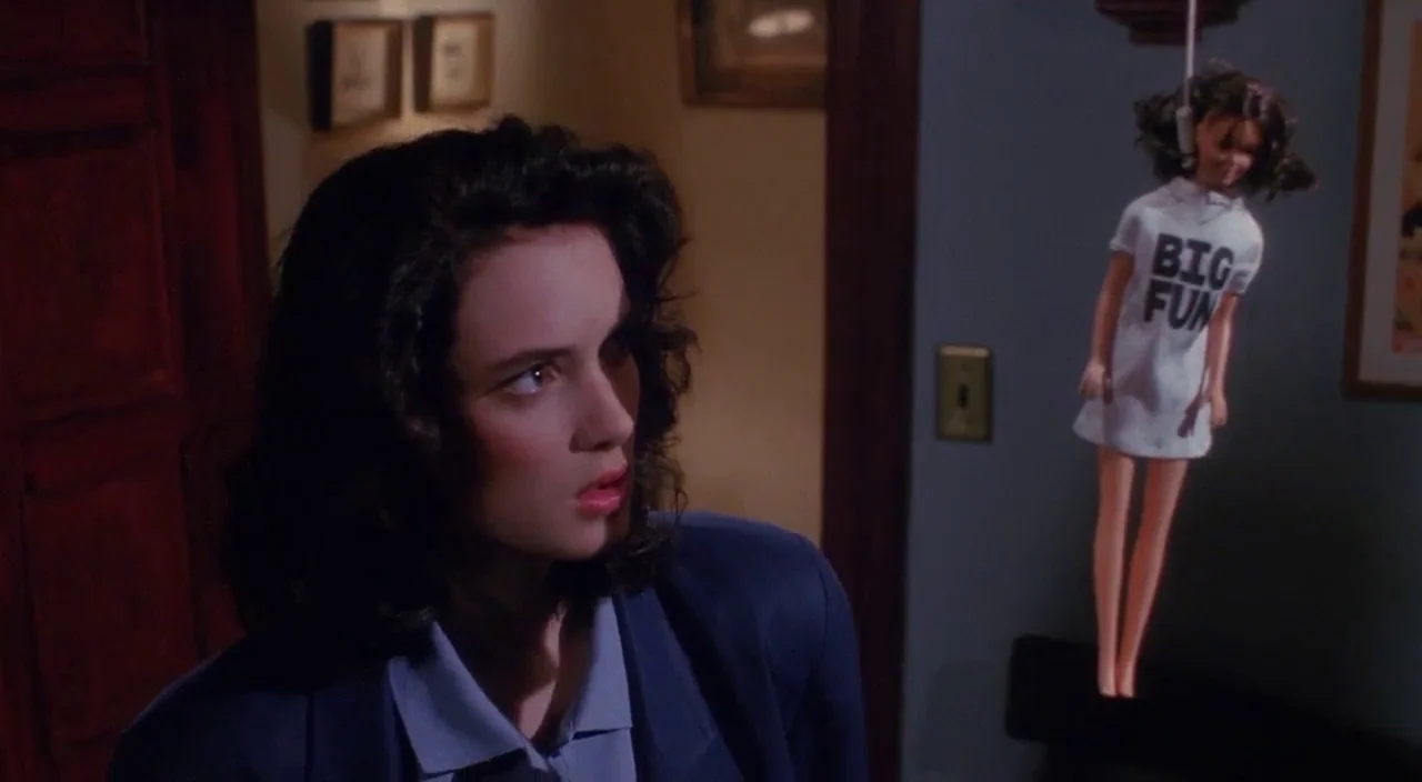 Heathers movie still: medium — A dimly lit indoor hallway with framed pictures on the wall and wooden trim. A small doll wearing a t-shirt hangs by…