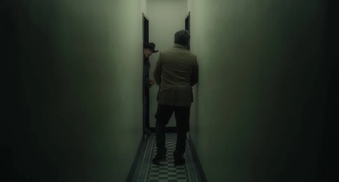 Inside Llewyn Davis (2013) movie still: fullbody — A dark narrow hallway features plain walls and a black and white checkerboard floor. One man in a…