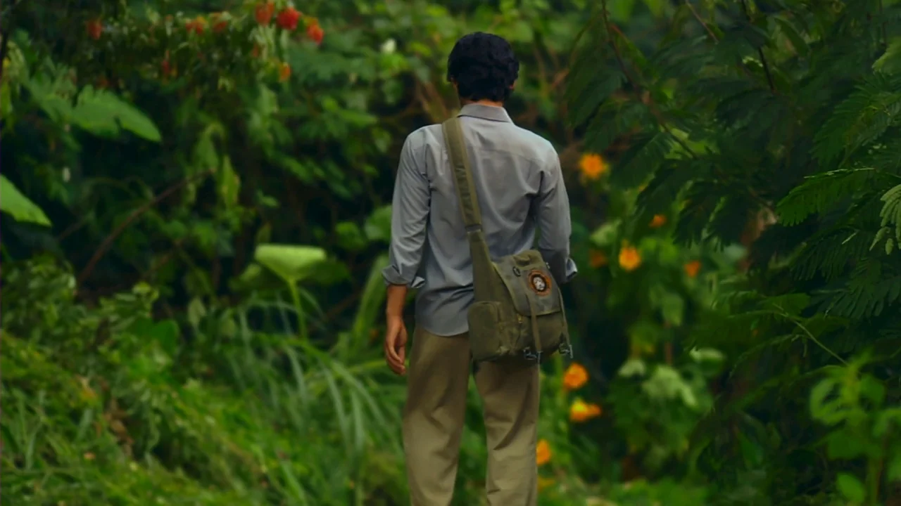 Merantau (2009) movie still: fullbody — A dense green jungle path is lined with thick foliage and yellow wildflowers. A man wearing a grey shirt and…