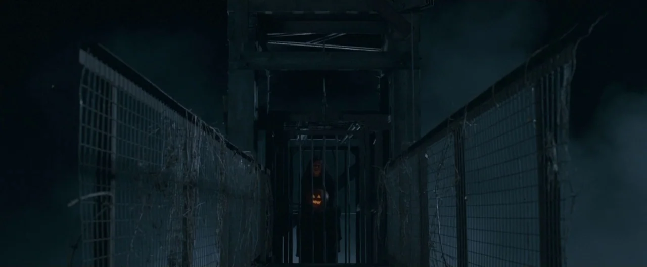 Trick ‘R Treat (2007) movie still: wide — A night scene shows a dark metal caged structure over a suspension bridge area. A figure stands within the…