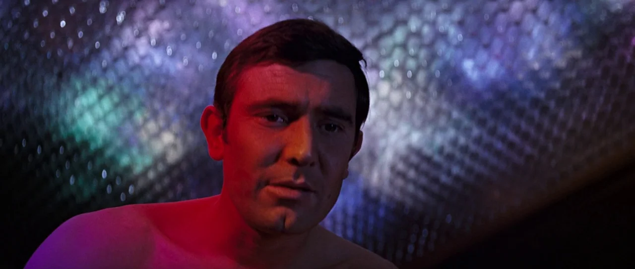 On Her Majesty’s Secret Service (1969) movie still: closeup — A man stands before a textured, metallic wall illuminated by shifting colored light.…
