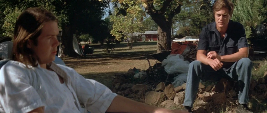 Shotgun Stories (2007) movie still: medium — A rural yard with trees and a scattered debris pile on the ground. Two young men sit in the shade, one…