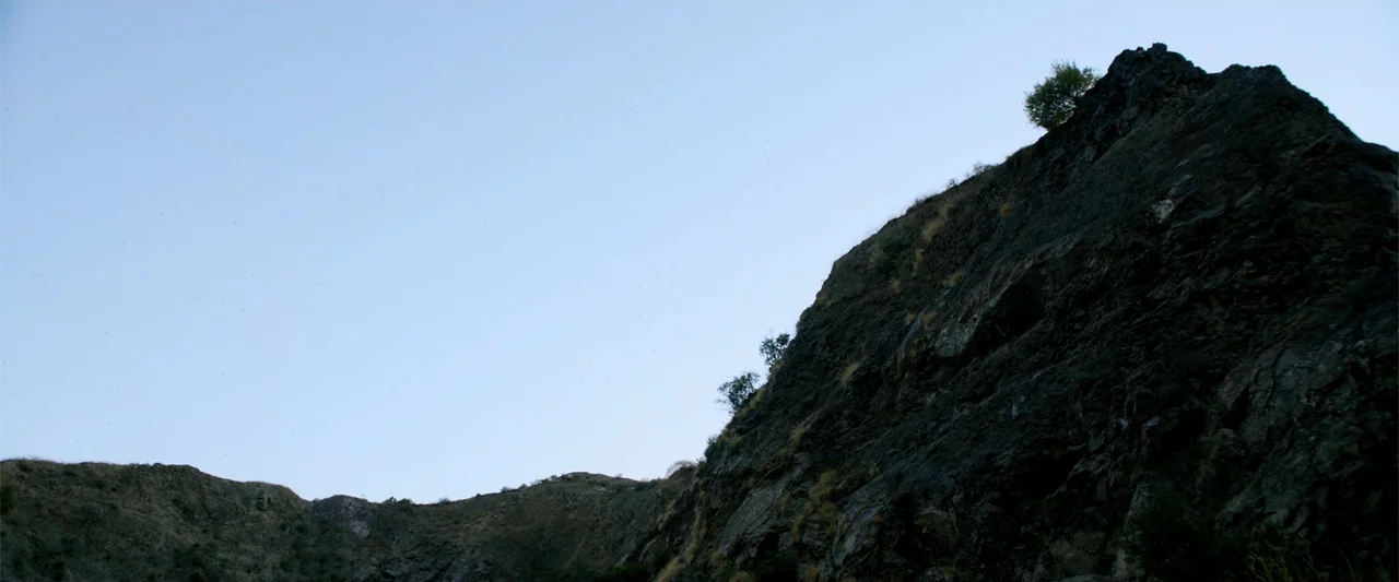 Nocturne (2020) movie still: establishing — A dark, jagged rock formation rises against a clear twilight sky. Small trees cling to the side of the…