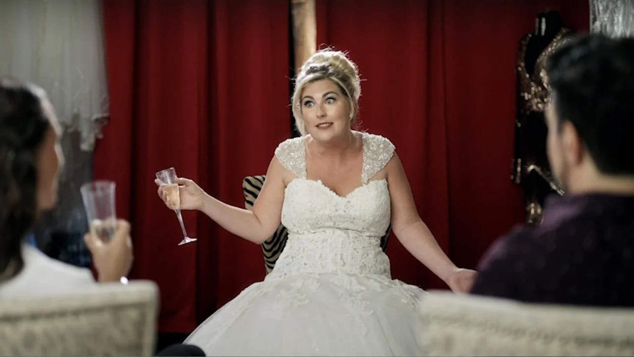 Ask Me to Dance (2022) movie still: medium — A woman in a bridal shop sits wearing a white wedding gown before red curtains. She holds a glass of…