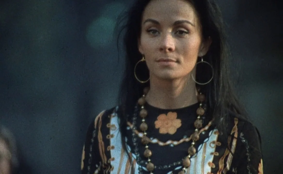 F for Fake (1973) movie still: medium — An outdoor setting provides a blurred dark backdrop behind a woman. The woman gazes forward while wearing…