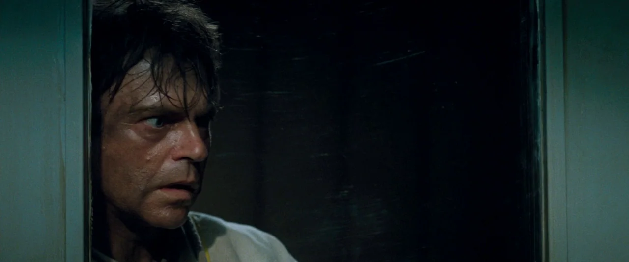 In The Mouth of Madness (1994) movie still: closeup — A man peers through a narrow vertical opening between a door and frame. Darkness occupies the…