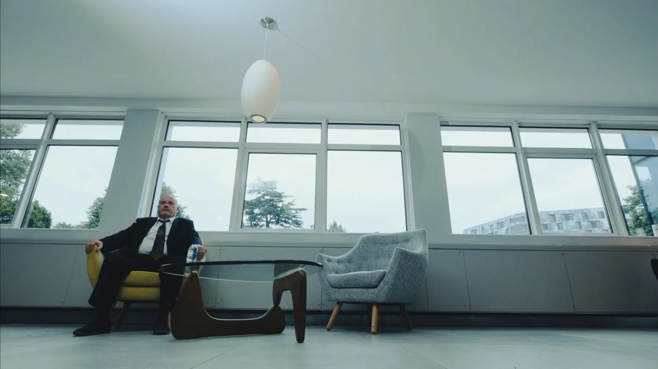 You Might Get Lost (2021) movie still: wide — A spacious room with large windows features a yellow chair and a glass table. A man in a suit sits in…