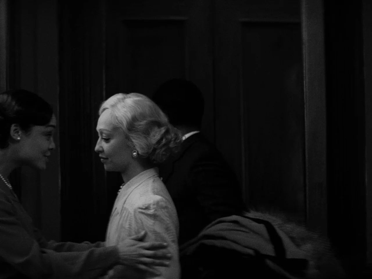 Passing (2021) movie still: medium — A dark hallway with shadowed double doors and architectural wood paneling in the background. Two women stand…