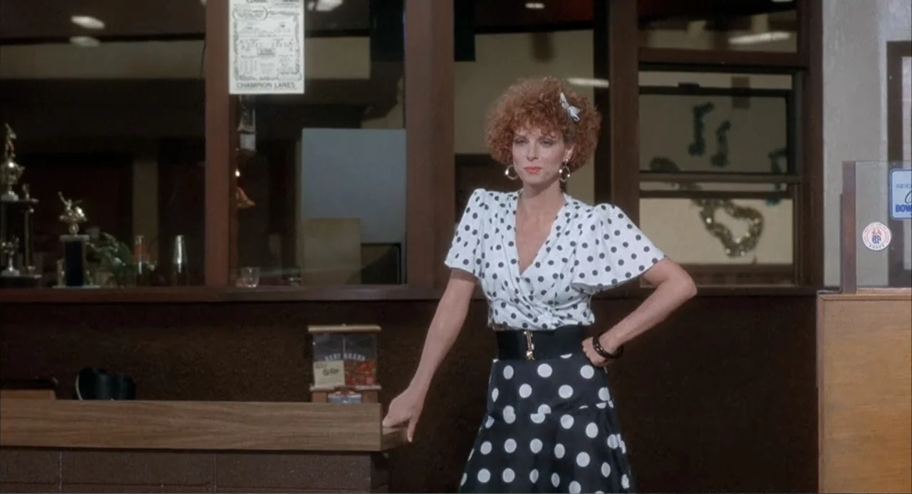 Overboard (1987) movie still: medium — A bowling alley interior features a wooden service counter and a dark windowed wall. A woman wearing a…